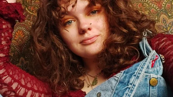 Daisy, a young woman with curly hair wearing denims, poses for a selfie.