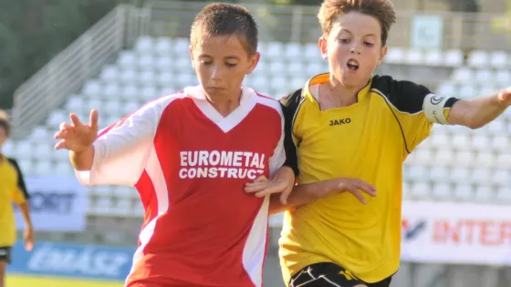 Two young boys wearing football kits running together on a football pitch. They are shoulder-barging each other and running towards the viewer.