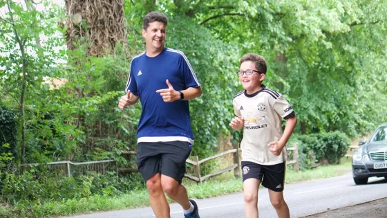 A young boy in glasses runs alongside his father on a tree-lined street, both smiling.