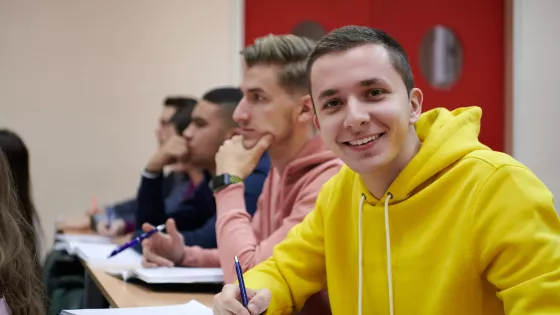 A university student in a yellow jumper smiles to camera as his peers look to the front of the class.