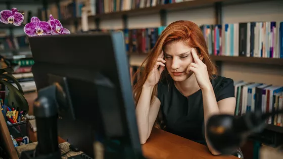 A young woman with red hair massages her temples in stress before a computer.