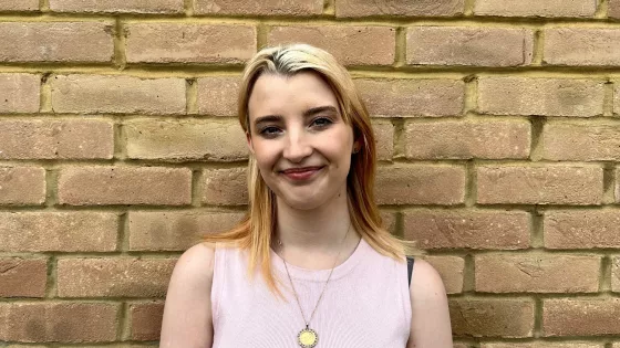 A young blonde woman, Bethany, stands against a brick wall.