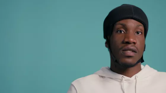 A young black boy in a wheelchair with a protective helmet against a blue backdrop wearing a white t-shirt