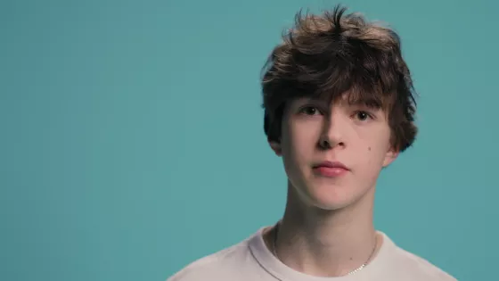 A young Caucasian boy stands against a blue backdrop wearing a white t-shirt