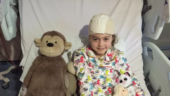 A young girl smiles from a hospital bed with a bandage on her head surrounded by teddy bears.