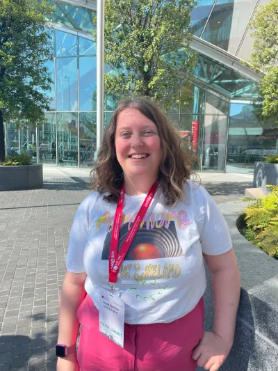 A young woman named Annie smiles to camera before a glass building on a sunny day.