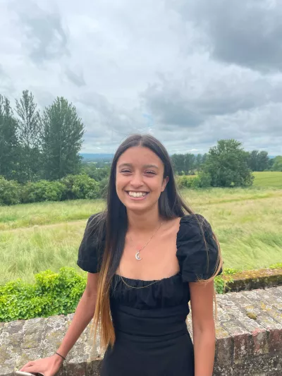 Jasmine, a young woman in a black t-shirt, smiles widely for a photograph before a grassy, tree-lined field.