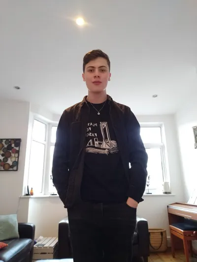 A young man in dark clothes stands for a photograph in a home.