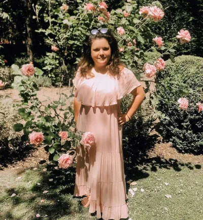 A young woman in a peach dress and sunglasses stands in a rose garden.