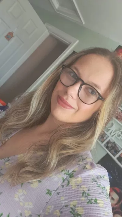 A young woman with long blonde hair and glasses, Jess, smiles in a selfie in her bedroom.