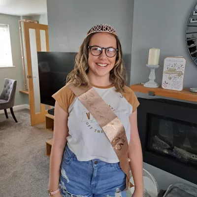 A young woman on her sixteenth birthday smiles while wearing a tiara and 'sweet sixteenth' sash.