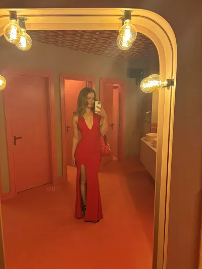 A young woman in a floor-length red dress poses in a mirror. 