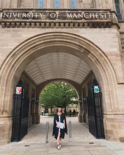 A young woman stands in a gown and cap under an arch at Manchester University at graduation.