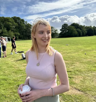 A young blonde woman, Bethany, holds a cup of coffee and smiles outside on a sunny day.