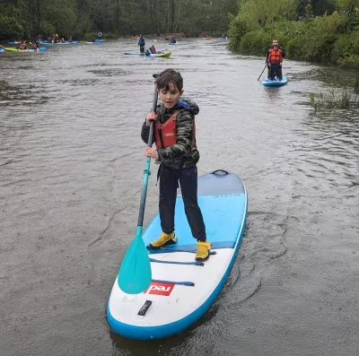 A young boy, Fin, paddleboards through a river in a wetsuit and life-jacket.