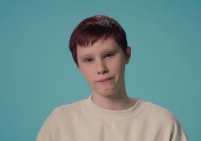 A young teenage white boy stands against a blue backdrop wearing a white t-shirt