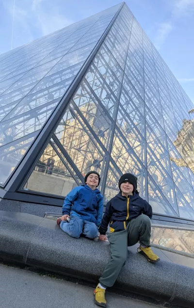 Two boys in winter clothing sit at the base of the Louvre pyramid.