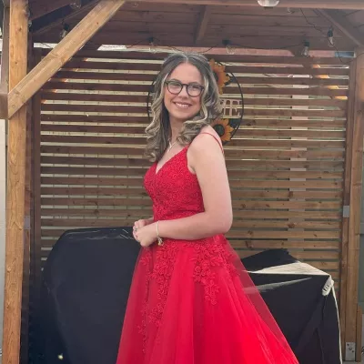 Jess, a young woman in glasses, smiles wearing a floor-length red ballgown.