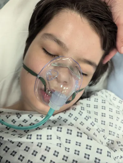 A young boy with a respirator lies with his eyes closed in a hospital bed.