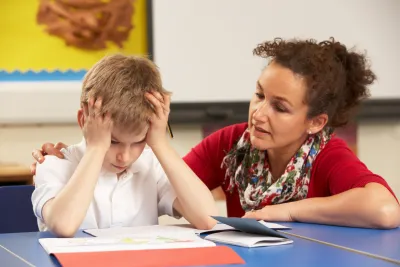 An educator speaks gently to a young student, who is holding his hair in frustration.