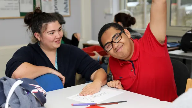 Male student in a red t-shirt, with a support worker, smiling with his hand raised.