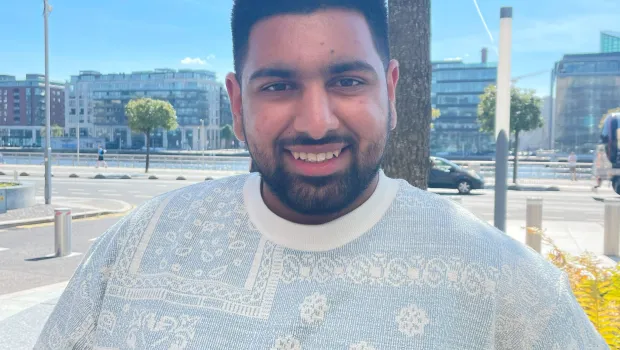 A bearded young man in a white t-shirt smiles into the camera on a summery day.