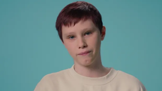 A young teenage white boy stands against a blue backdrop wearing a white t-shirt