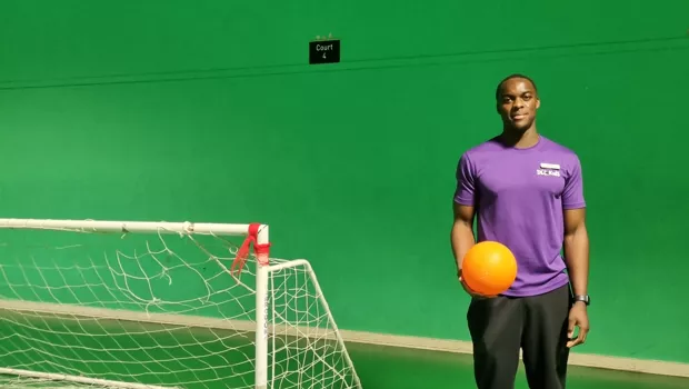 Renell, a young man in a purple t-shirt, stands with a football beside a goal against a green screen.
