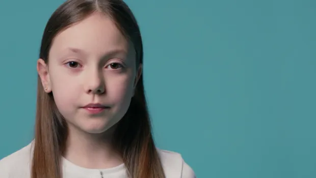 A young Caucasian girl stands against a blue backdrop wearing a white t-shirt