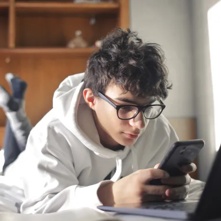 Young boy lying on his bed looking at his phone and laptop