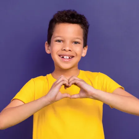 Young boy smiling in a yellow t-shirt holding a hand heart against a purple backdrop.