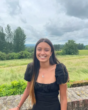 Jasmine, a young woman in a black t-shirt, smiles widely for a photograph before a grassy, tree-lined field.