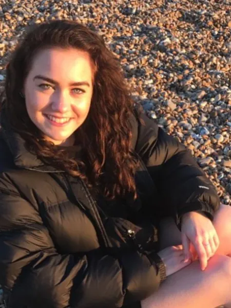 A young woman sits on a pebble beach, smiling for the camera.