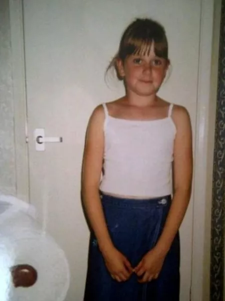 A young girl in a white vest smiles slightly in a posed childhood photograph.