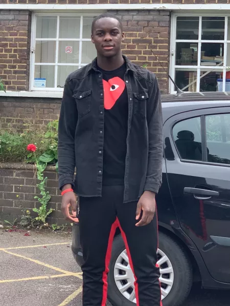 A young man wearing dark clothes with orange accents stands in front of a house.