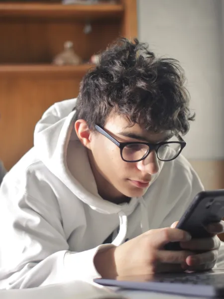 Young boy lying on his bed looking at his phone and laptop