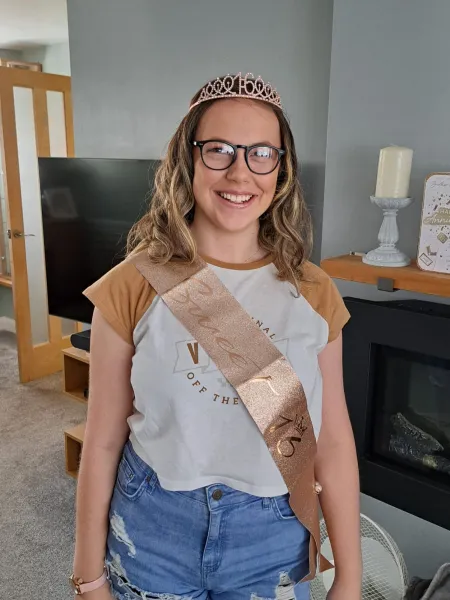 A young woman on her sixteenth birthday smiles while wearing a tiara and 'sweet sixteenth' sash.