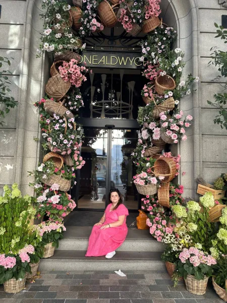 A young woman sits on the step of a doorway, surrounded by baskets of pink flowers.