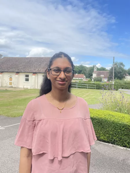 A young woman, Tee, smiles in a rural area, wearing glasses and a peach blouse.