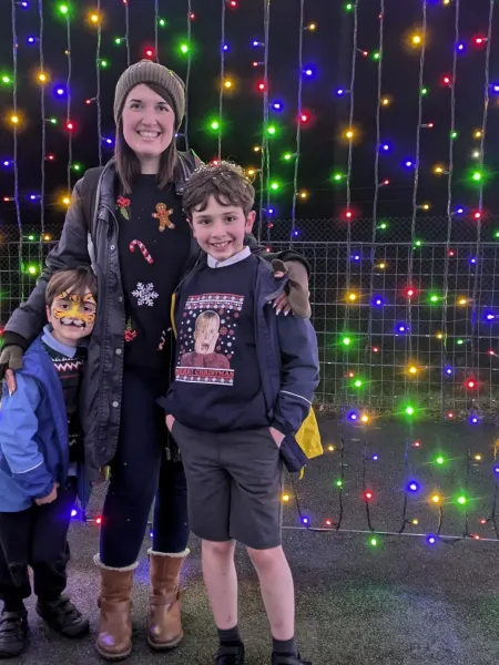 A mother and her two sons smile at Christmas time in front of a wall of fairy lights.