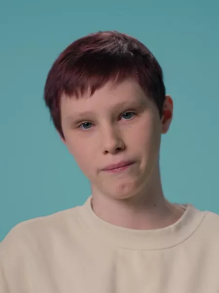 A young teenage white boy stands against a blue backdrop wearing a white t-shirt