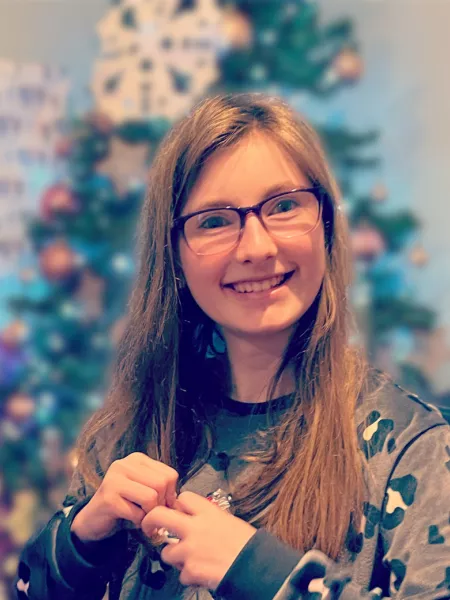 A young woman in glasses and pyjamas smiles before a Christmas tree.