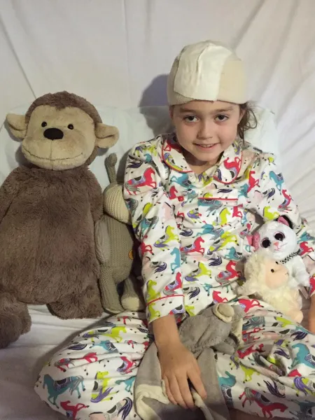 A young girl with a surgery bandage on her head sits with her stuffed teddies.