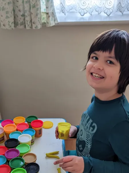 A young boy smiles at a desk laden with colourful play doughs.