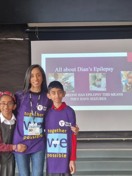 A mother stands with her two children in Young Epilepsy t-shirts in front of a screen.