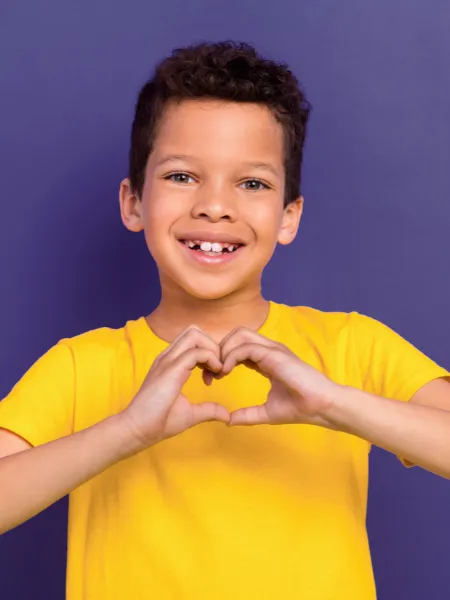 Young boy smiling in a yellow t-shirt holding a hand heart against a purple backdrop.